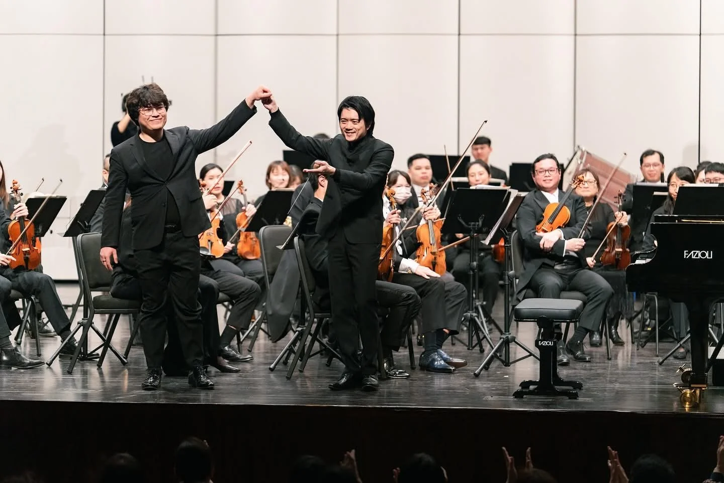 Two men in black suits smiling and holding hands in front of an orchestra on stage, with the orchestra members holding violins, violas, and a piano visible.