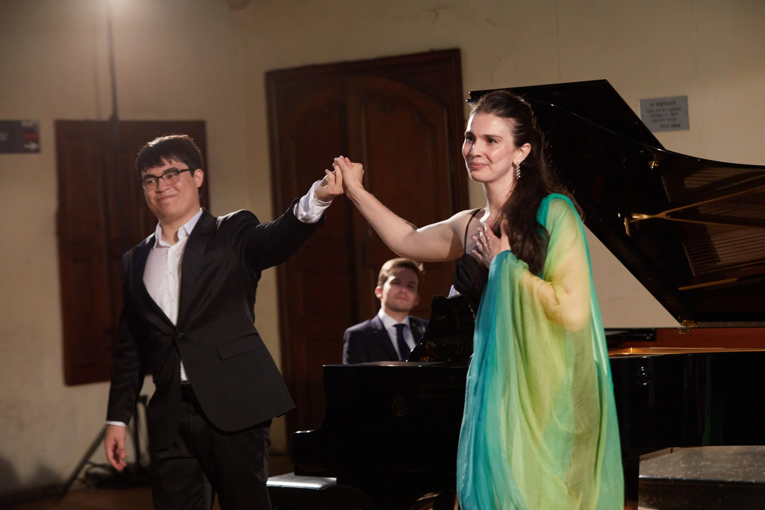 A man and woman on stage, holding hands and smiling, after a performance. The woman is wearing a colorful dress and the man is wearing a black suit. There is a pianist in the background.
