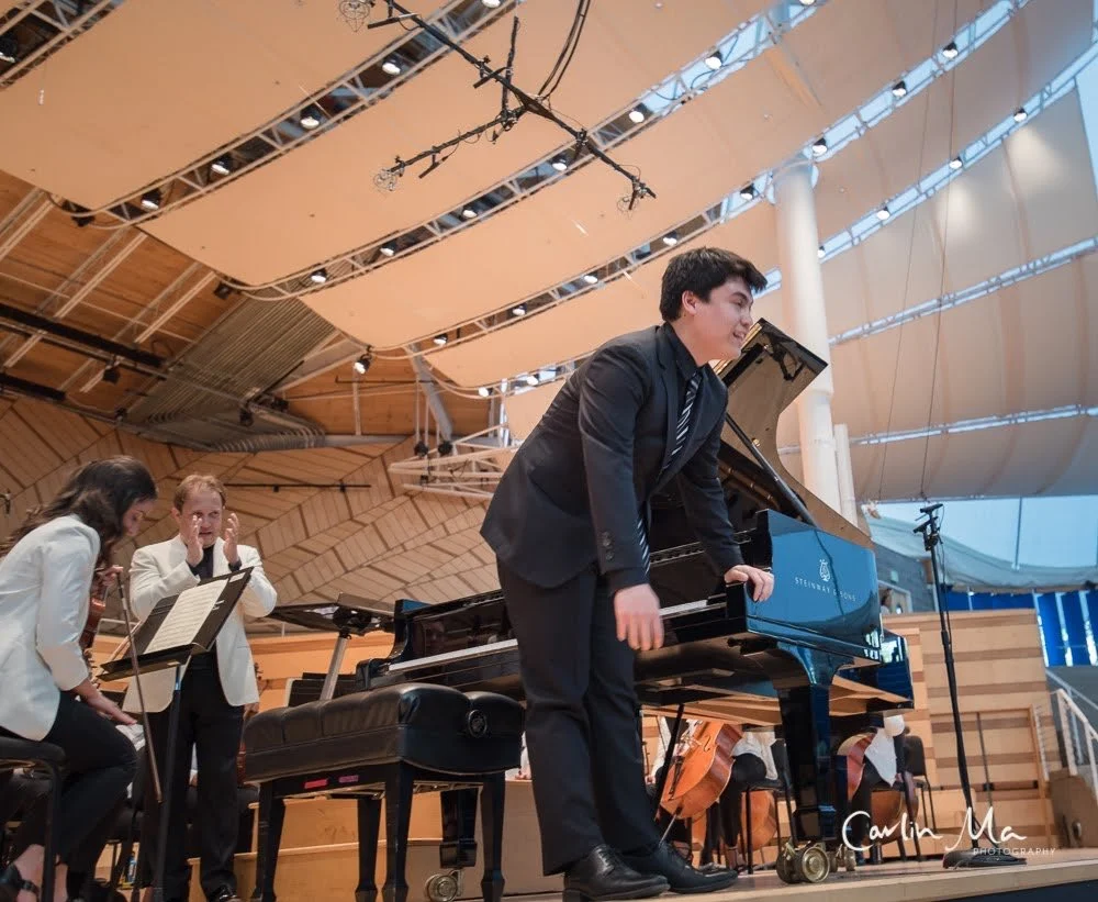 A young man in a suit bowing at a concert stage with a grand piano, with two women reacting with surprise and applause, in a large hall with wood-paneled ceiling.