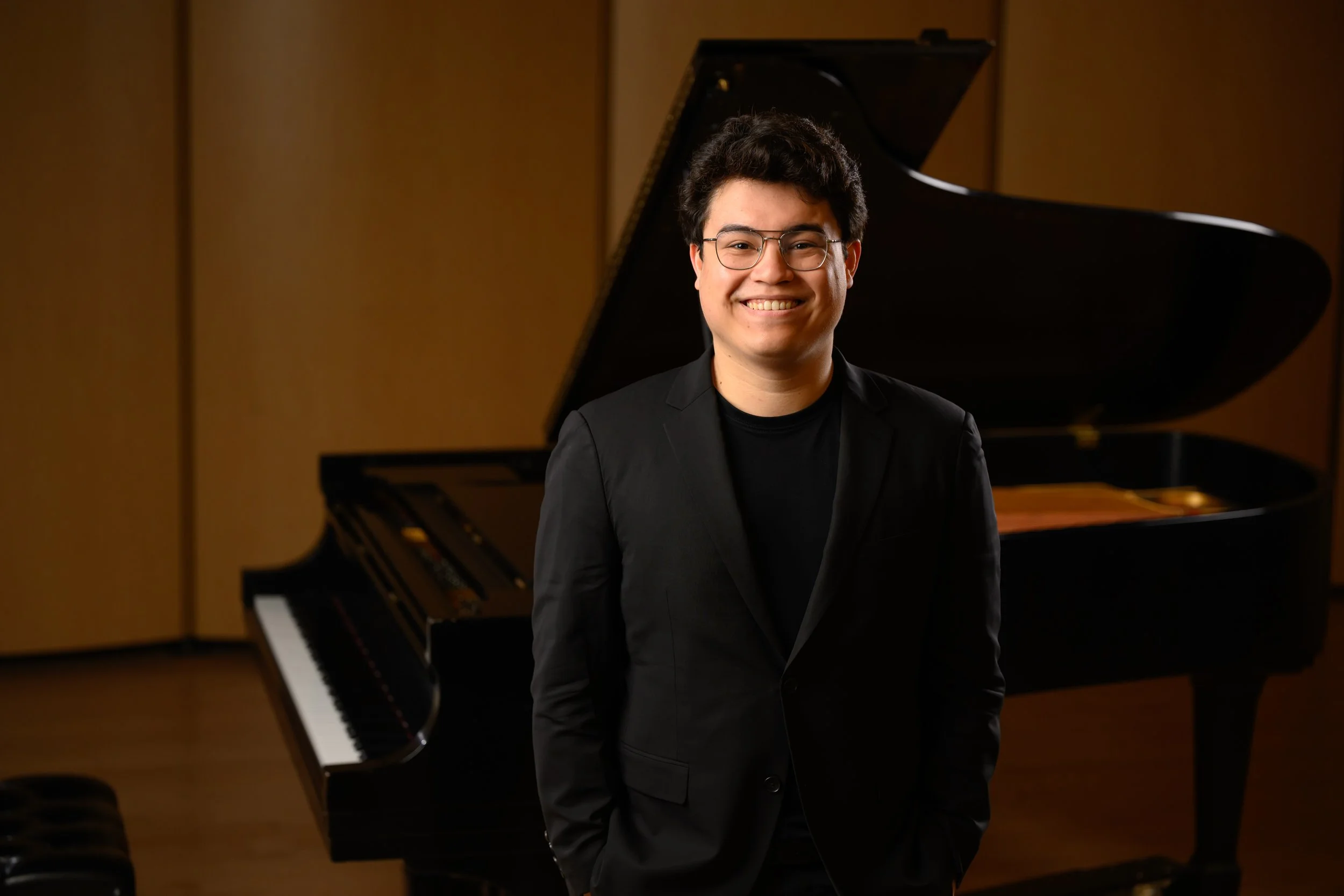A smiling man wearing glasses and a black suit standing in front of a grand piano in a music studio or concert hall.