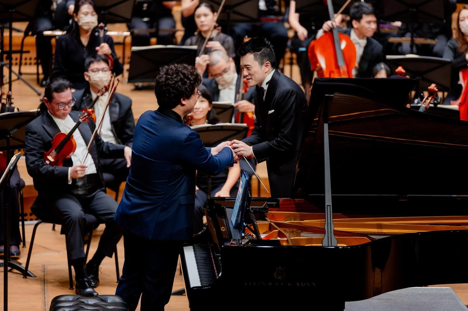 A man in a blue suit and a man in a black tuxedo are shaking hands on stage during a concert, surrounded by an orchestra with string and wind musicians.