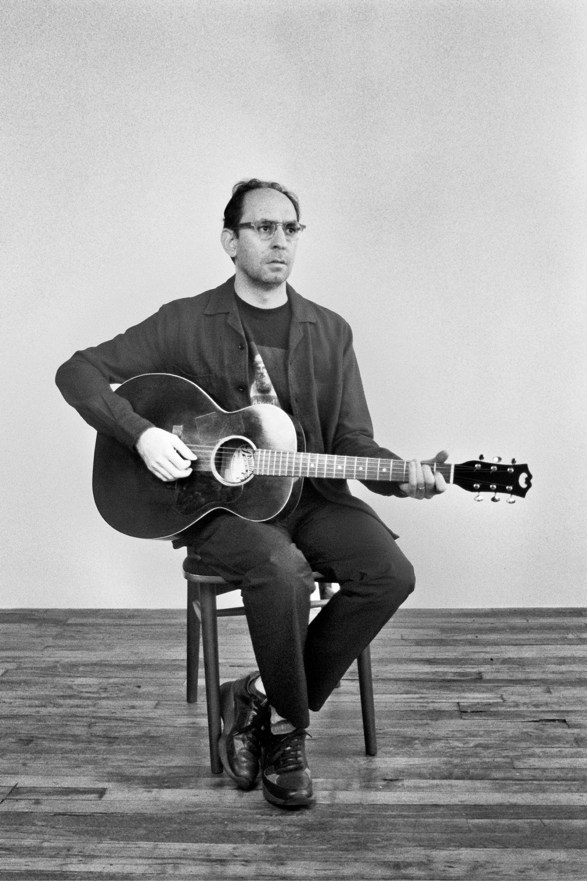Black and white image of Rami Gabriel. A man with glasses, wearing a jacket and a graphic t-shirt, sitting on a chair and playing an acoustic guitar on a wooden floor with a plain wall background.