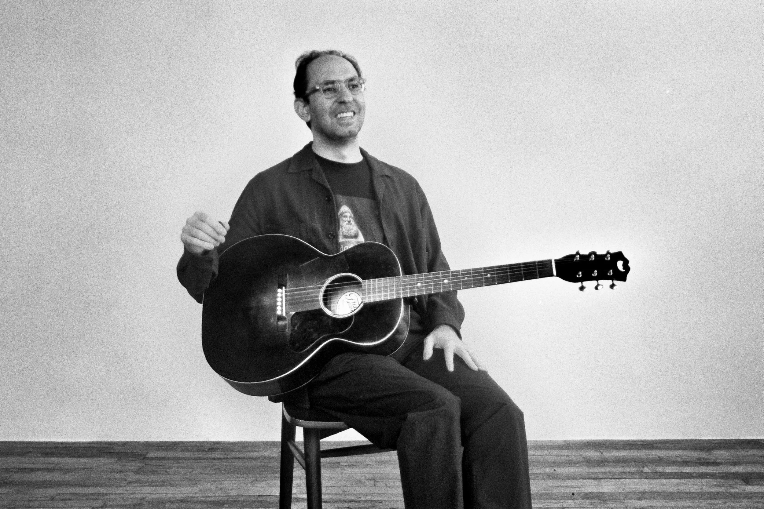 Black and white image of Rami Gabriel. A man sitting on a stool playing an acoustic guitar, smiling, in a plain room with wooden floors.