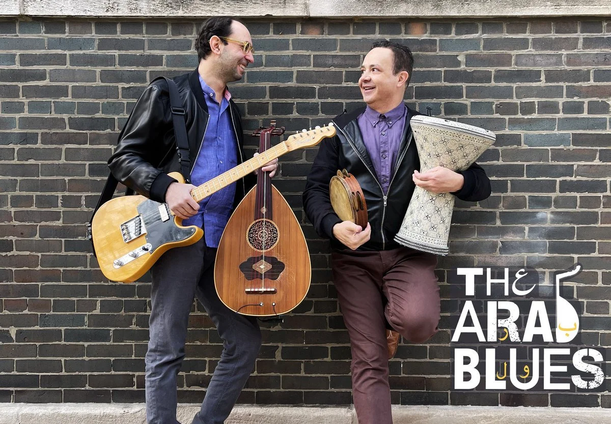 Album cover for "The Arab Blues" by The Arab Blues. Two men standing against a brick wall, holding musical instruments (oud, electric guitar, darbuka, and riq) and smiling at each other.