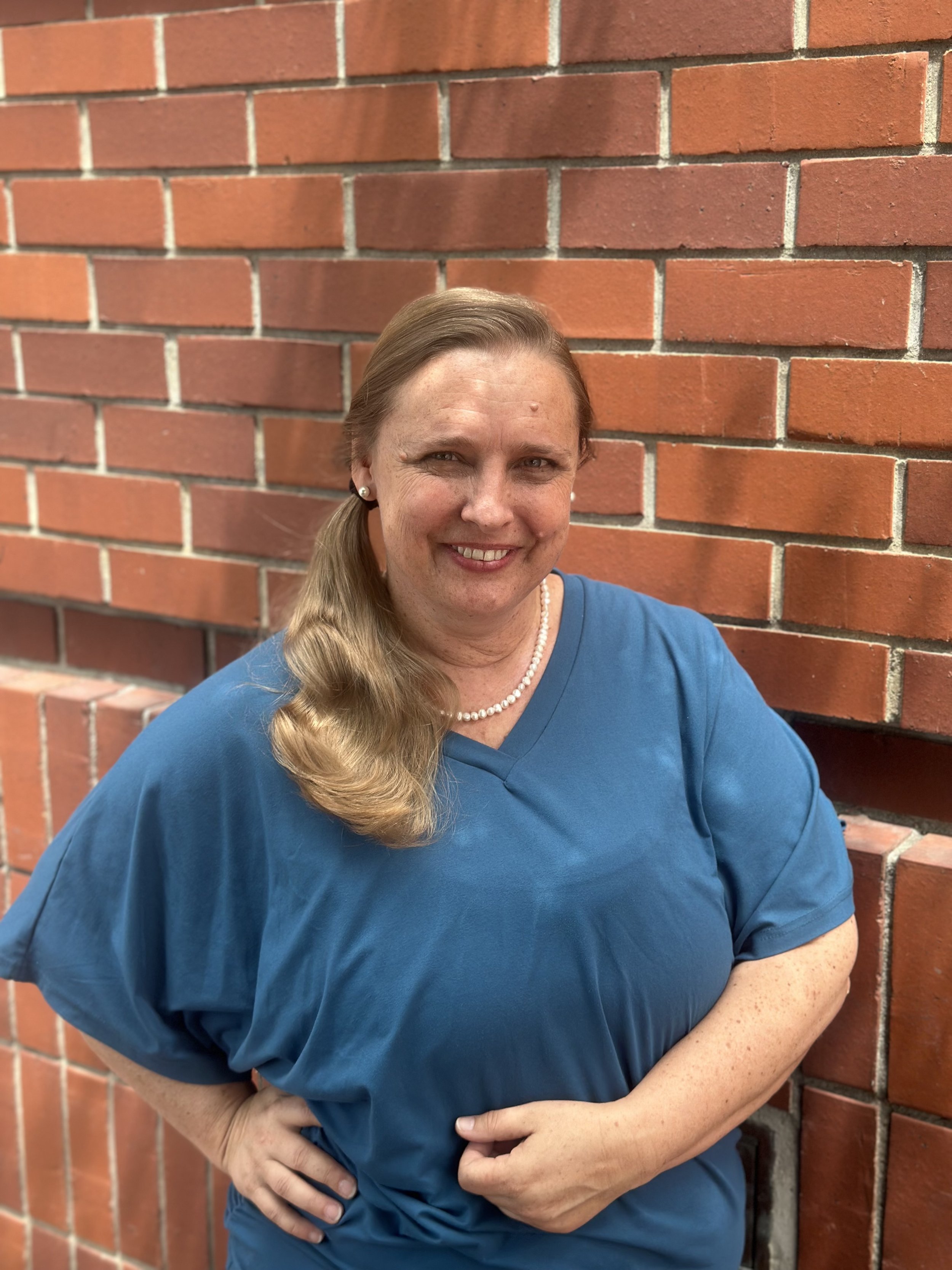 A woman in blue top with pearl necklace, standing against a red brick wall, smiling with arms crossed.