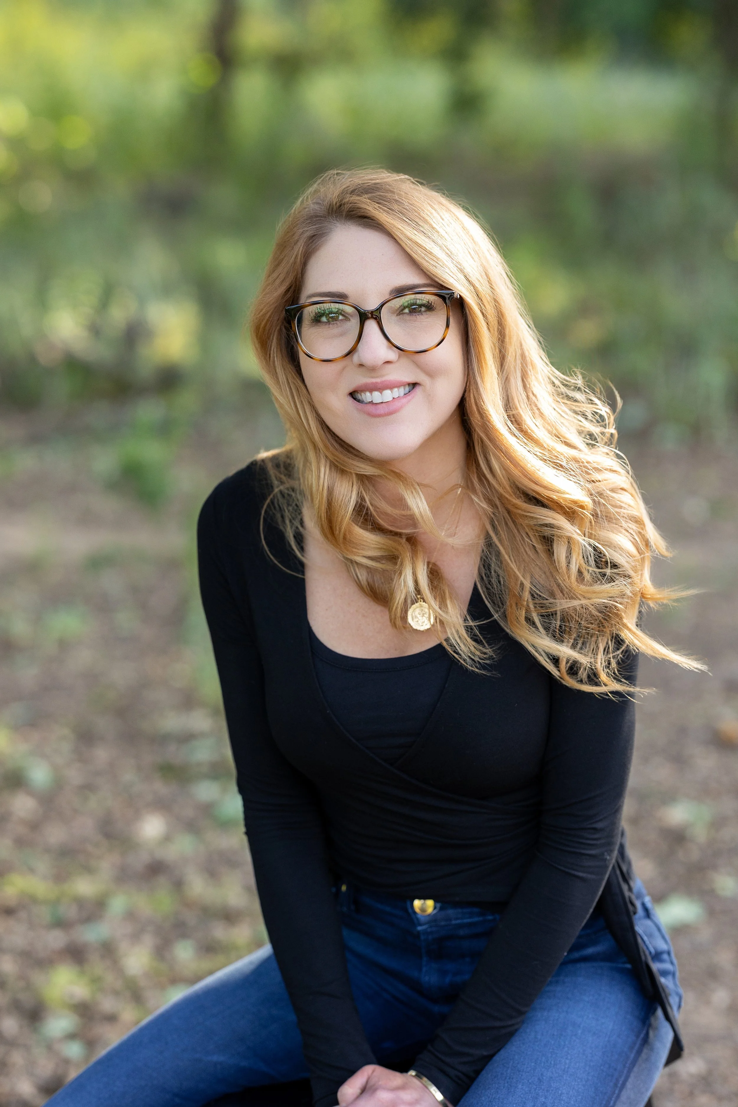 A woman with red hair and glasses smiling outdoors, wearing a black long-sleeve top and blue jeans, sitting on a rock with greenery in the background.