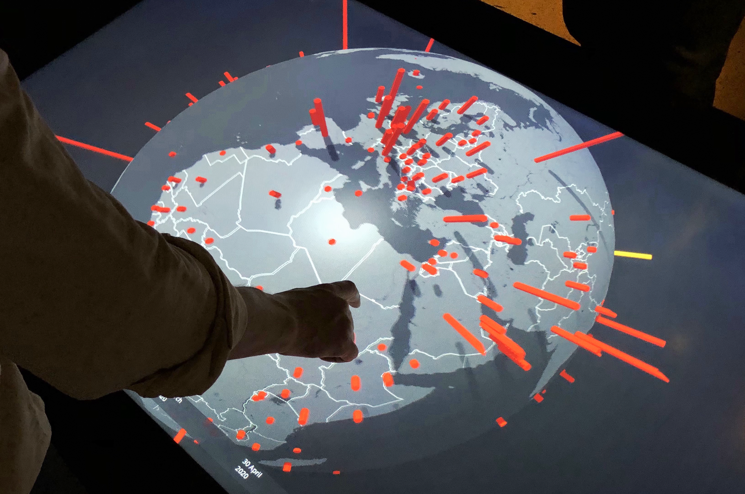 A person pointing at a large digital map displayed on a circular surface, showing a global map with various red 3D markers representing data points, with a focus on Europe and surrounding areas.