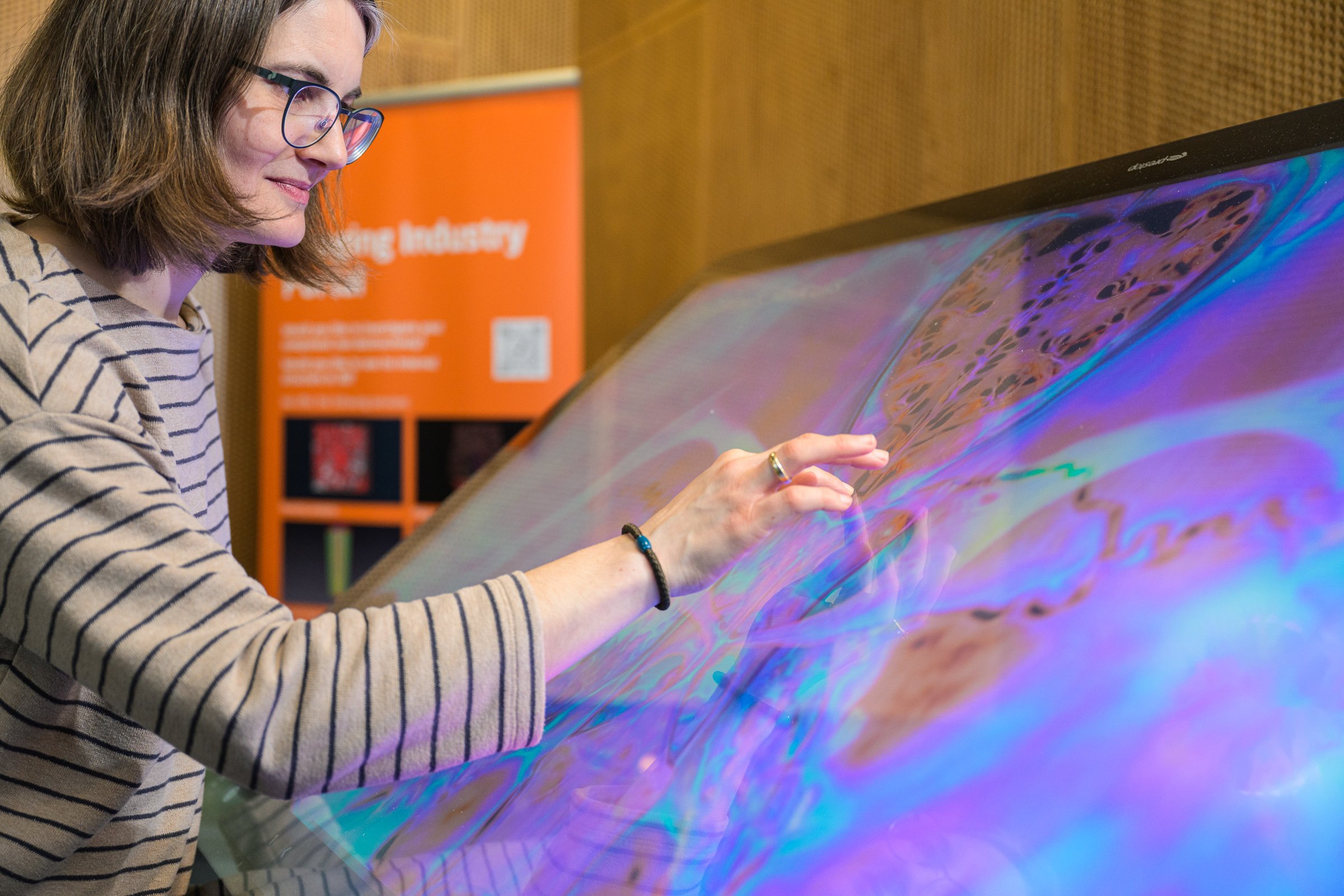 A woman with glasses and a striped shirt is touching a large, colorful, holographic touchscreen display with her finger.