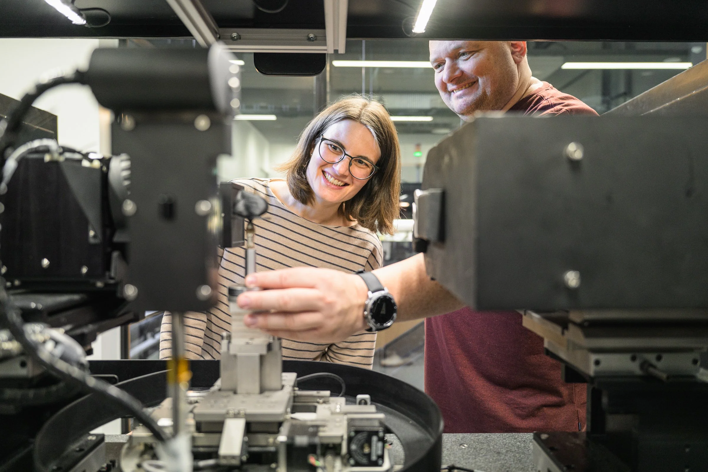 Two people working together on machinery in a lab or workshop environment, with various technical equipment around them.