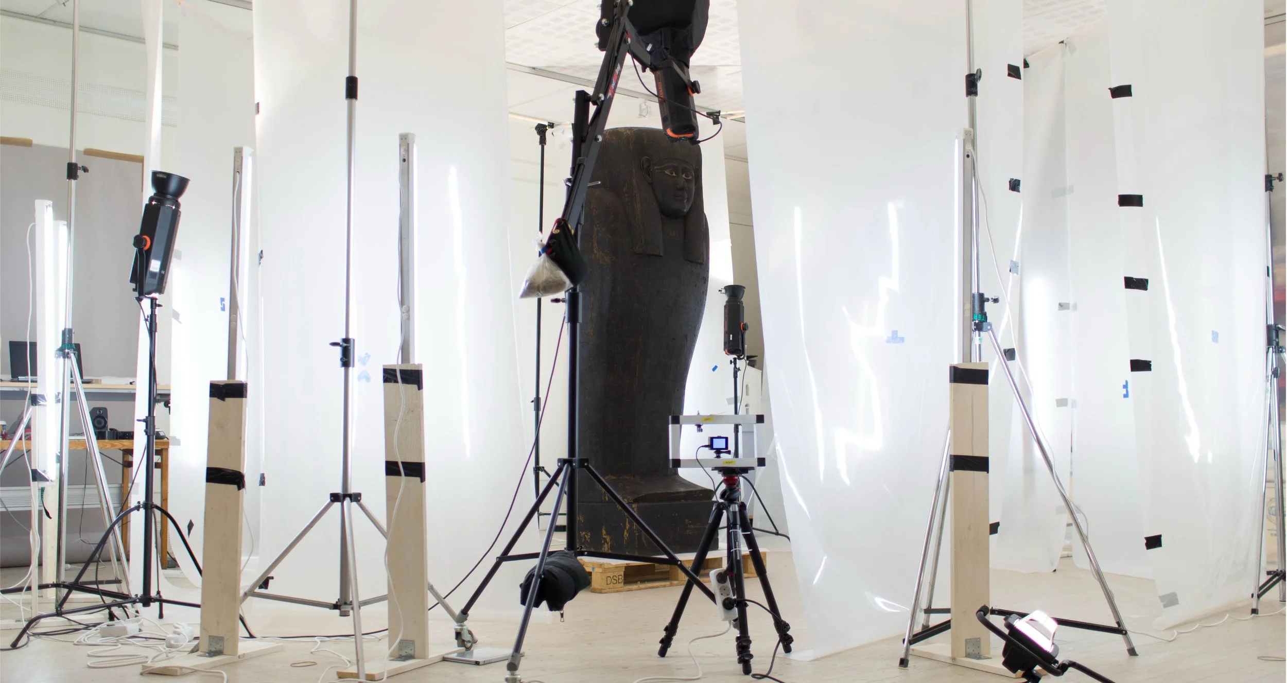 A photo of an art or archaeological exhibit with a large black artifact that appears to be a carved stone or sculpture of a pharaoh or figure with a headdress, set between white photographic screens and surrounded by various photography and lighting equipment.