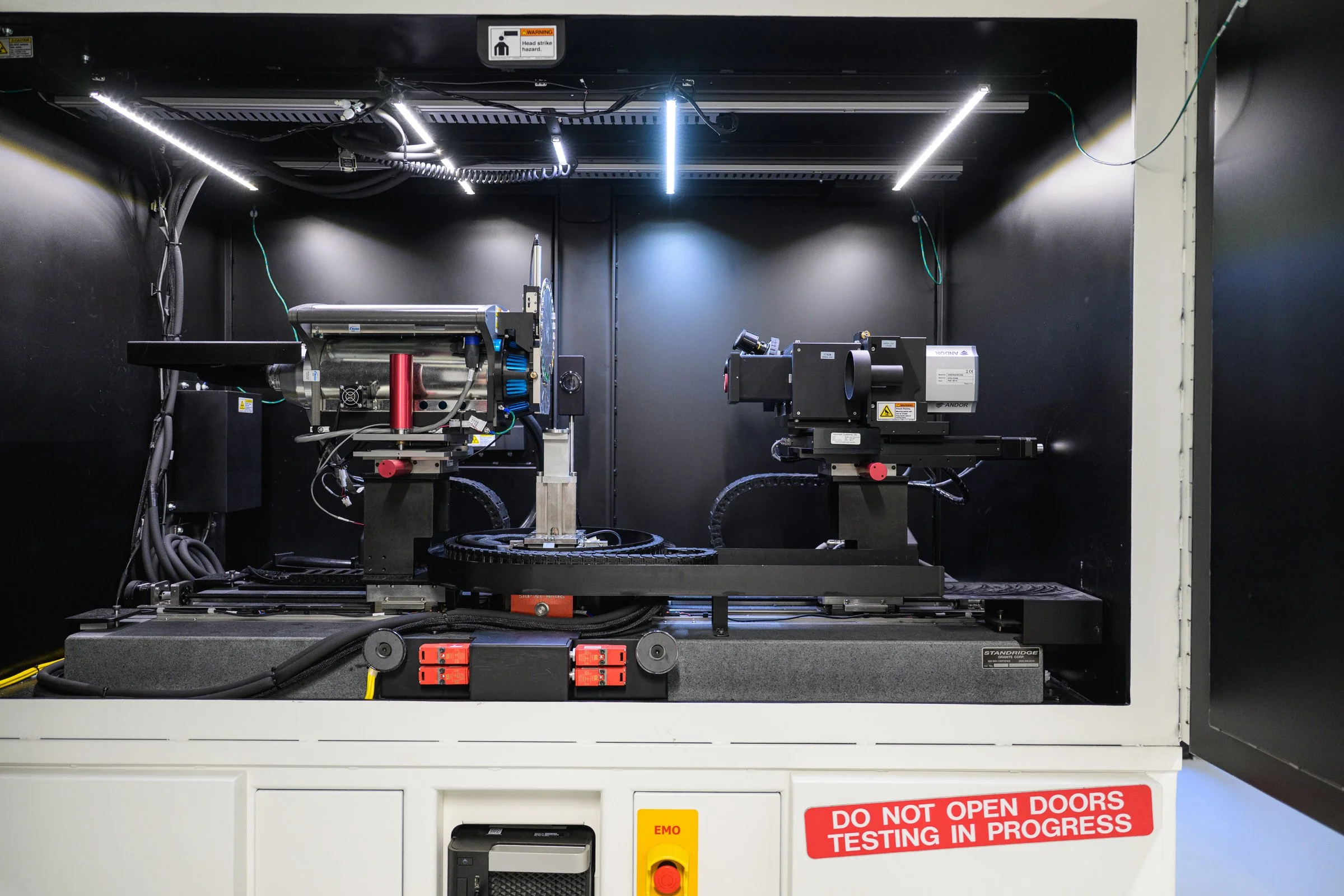 Inside a laboratory or testing facility, there are two advanced machines with electronic components and wiring, surrounded by safety signs including a warning about head strike hazard and a red label indicating testing in progress.