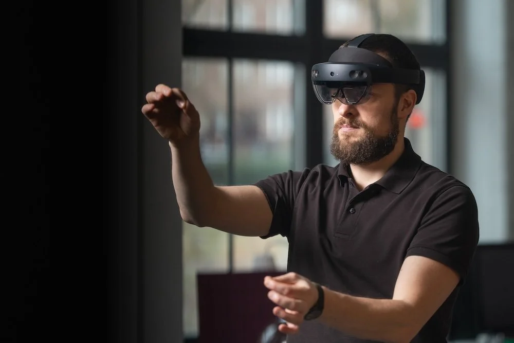 Man wearing a virtual reality headset, in a black polo shirt, inside a room with large windows.