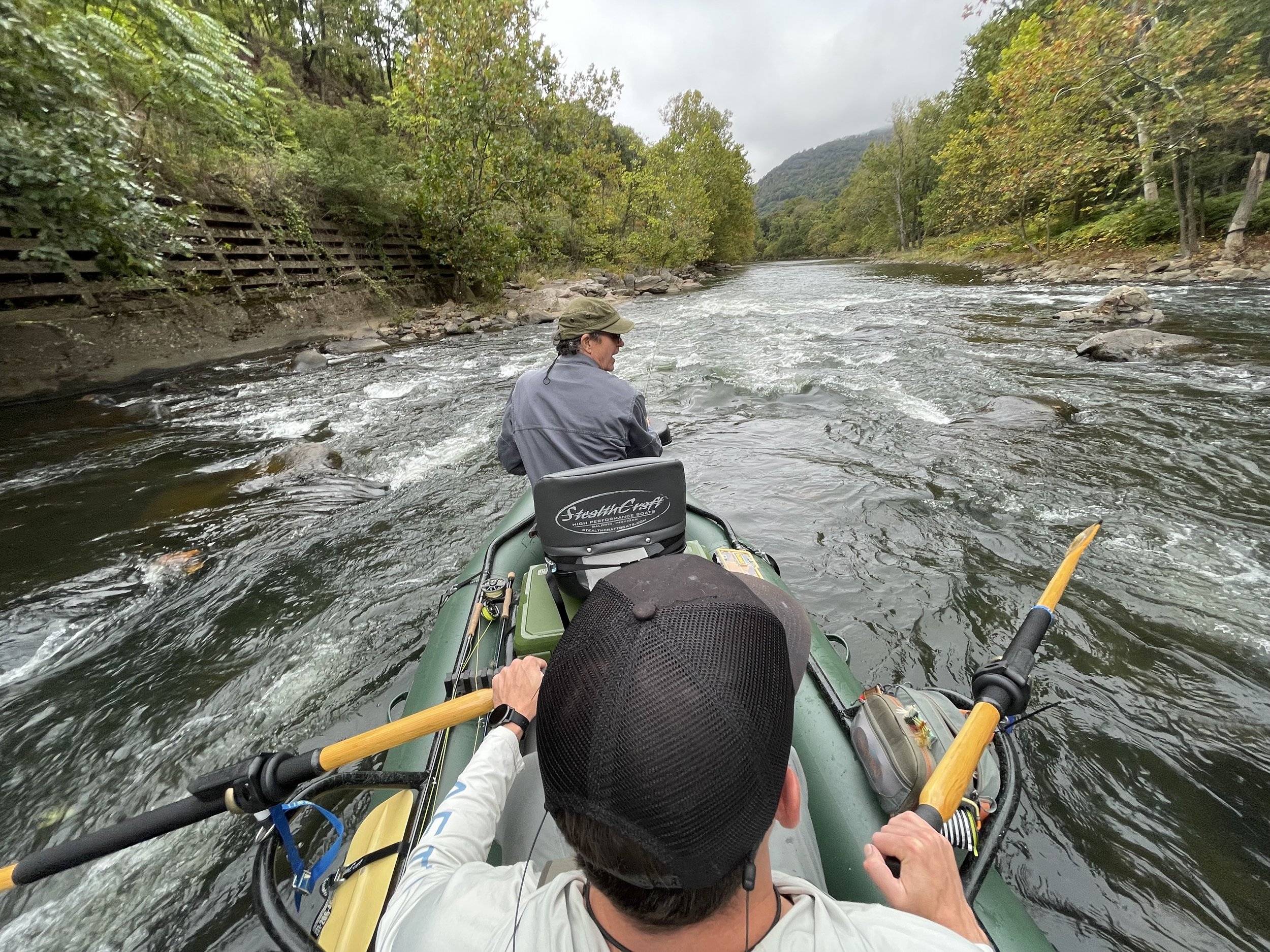 Two people fishing on a river in a green inflatable boat, surrounded by trees and hills, on a cloudy day.
