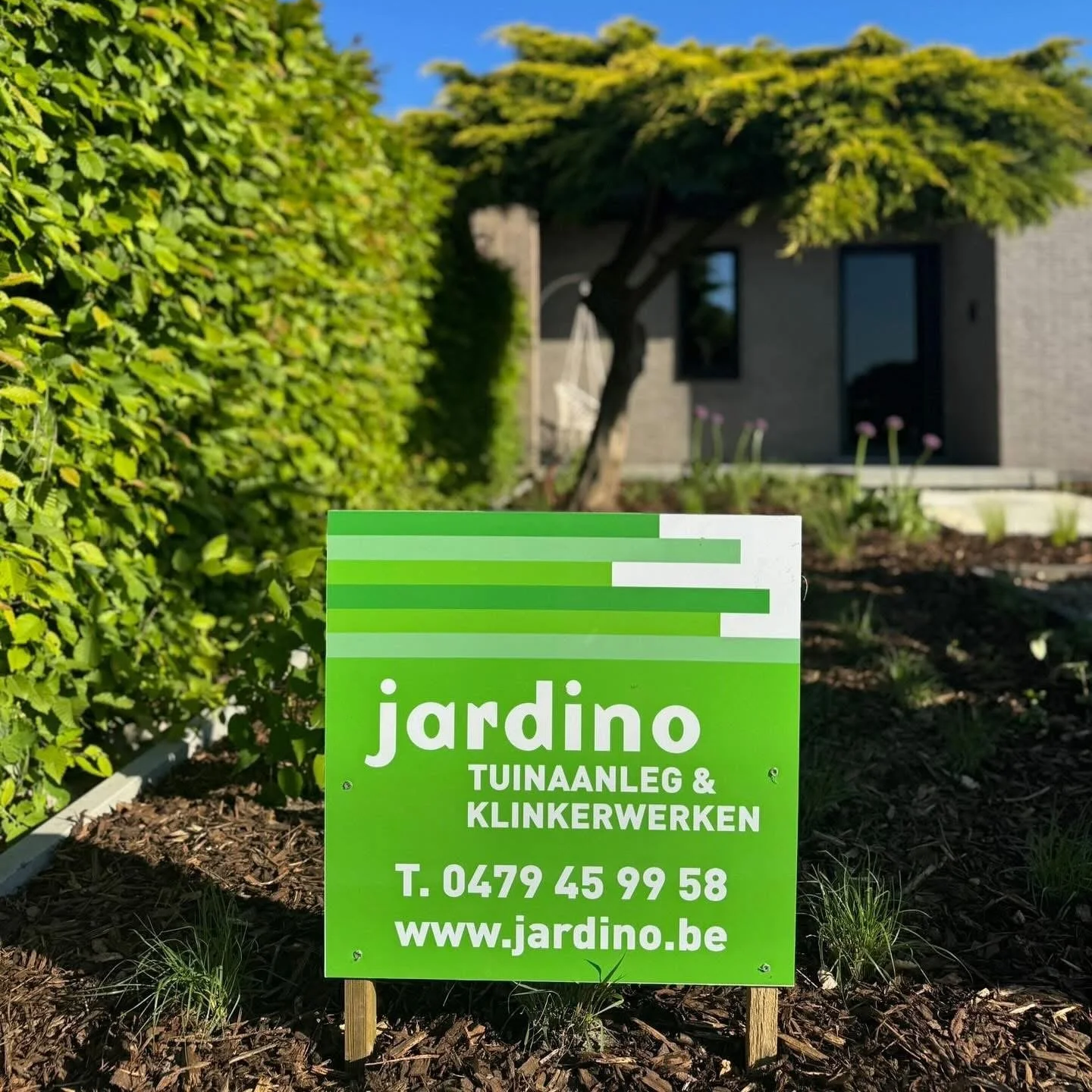 Groen tuinschapsbord met contactgegevens en website, geplaatst in een tuin met planten en een huis op de achtergrond.