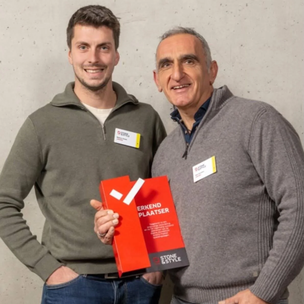 Twee mannen staan naast elkaar, één houdt een rode doos met witte tekst. Ze dragen allebei name tags en glimlachen. De achtergrond is een eenvoudige betonnen muur.