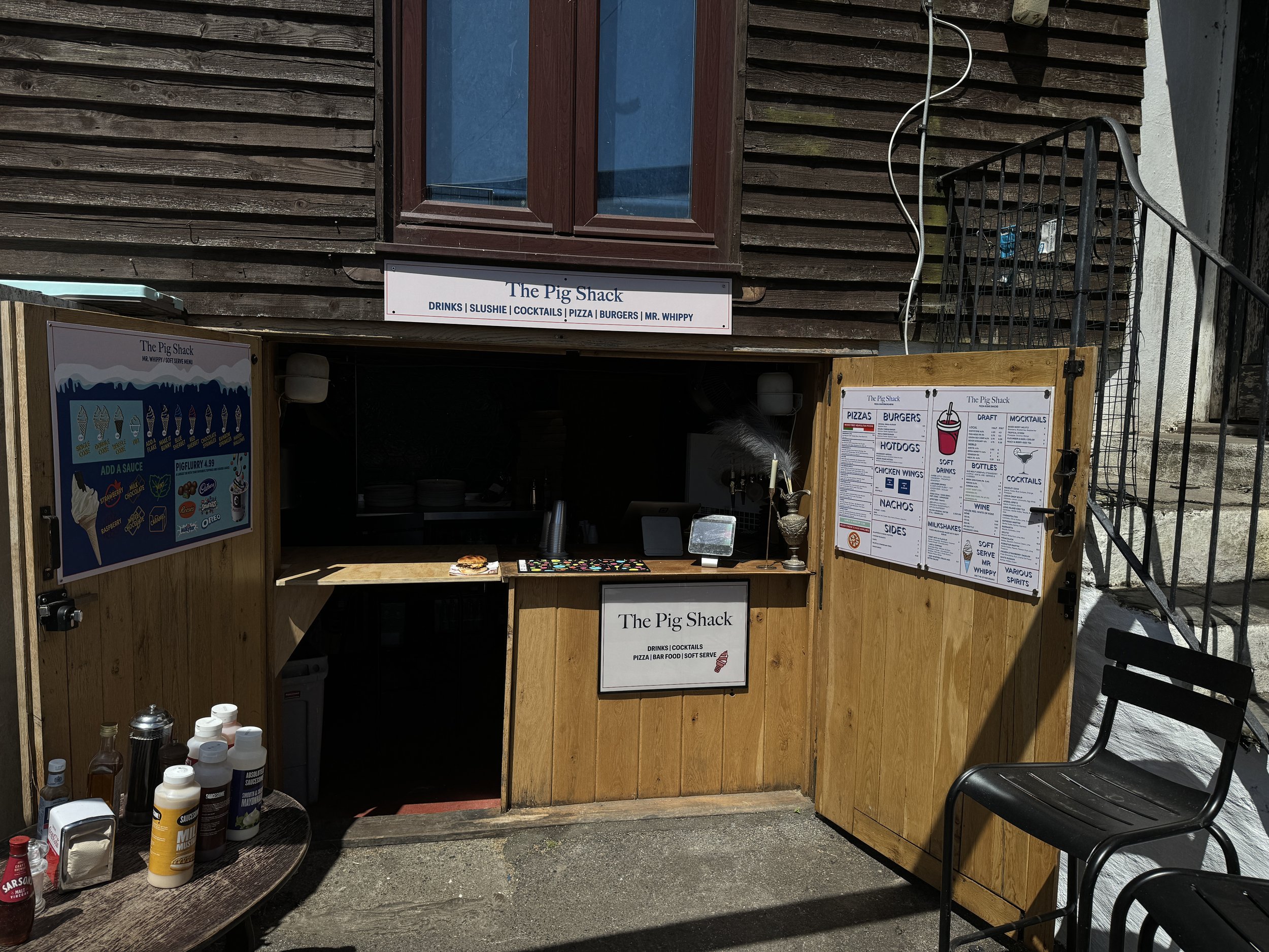 Small outdoor food stand called 'The Pig Shack' with menu boards displaying pizzas, burgers, hot dogs, drinks, and more. Contains condiments on a table, a black chair, and steps leading to a building. Wooden exterior with a sign above reading 'The Pig Shack' and additional signage.