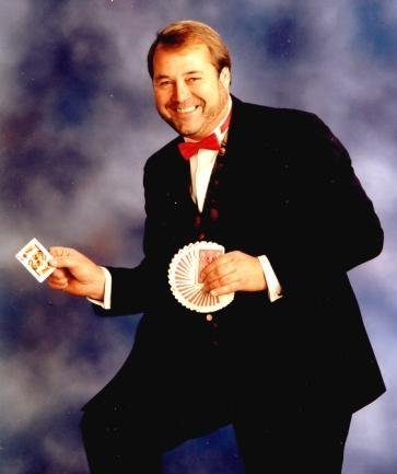 A man in a tuxedo with a red bow tie holding playing cards and a fan of playing cards, smiling at the camera.