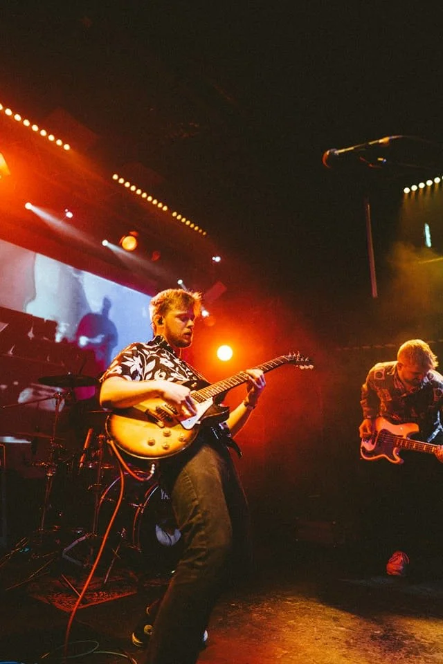 Two musicians playing electric guitars on stage under colorful lights, with a large screen in the background displaying abstract visuals.