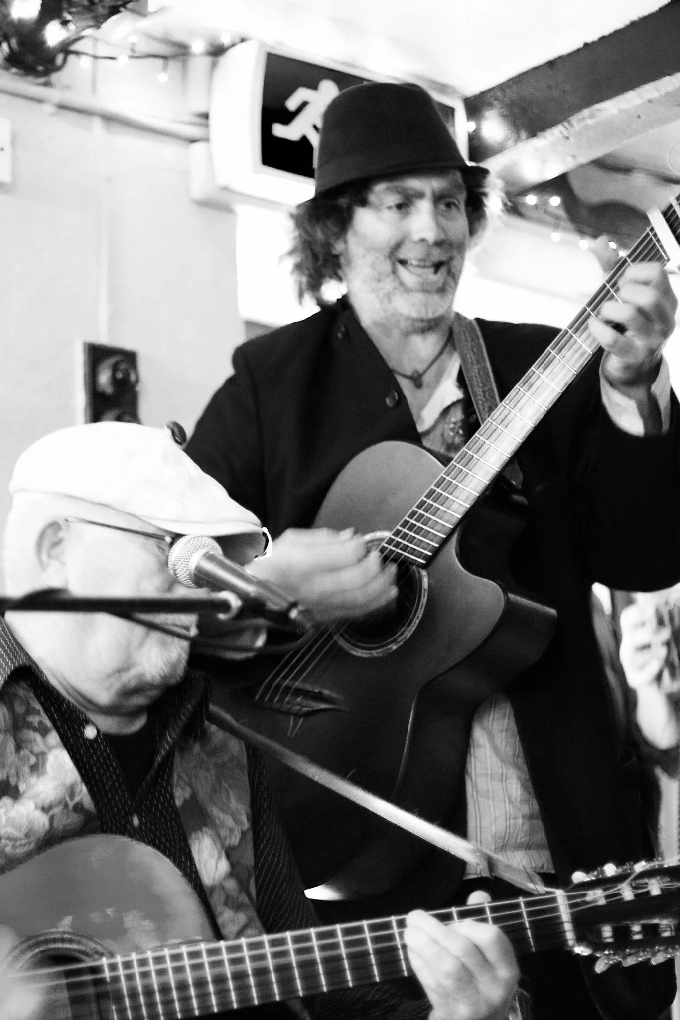 Two musicians playing guitars; one is singing into a microphone with a dark hat, and the other is wearing glasses and a cap. They are performing in a small, cozy setting, possibly a bar or cafe.