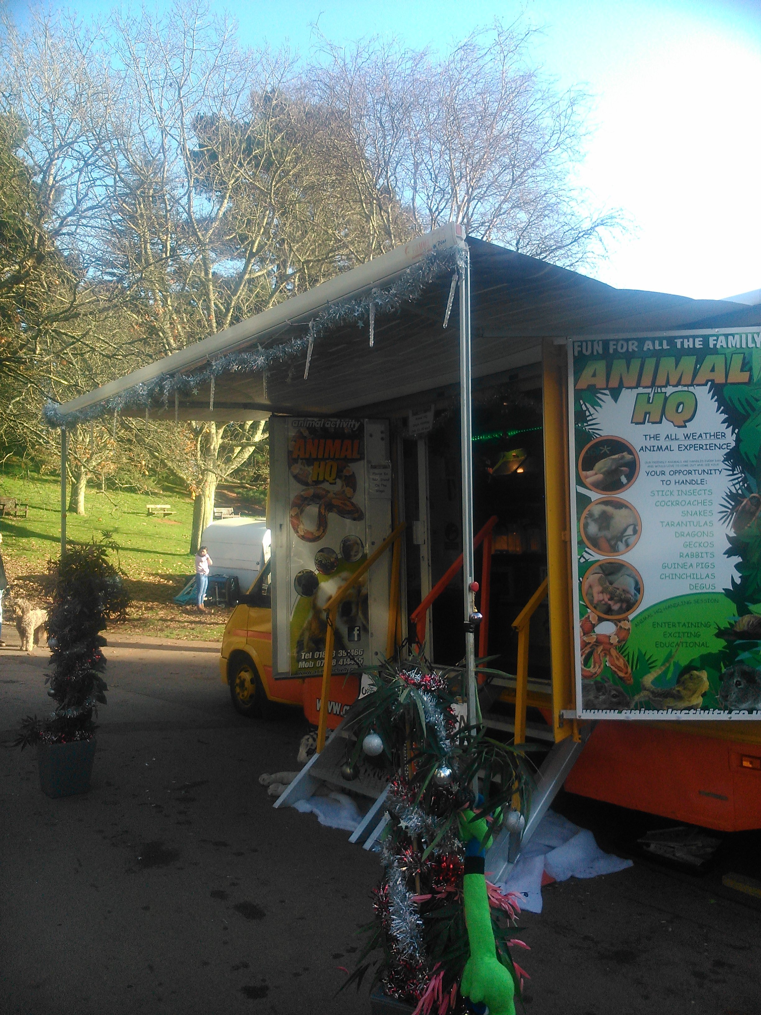 A small yellow carnival truck decorated with tinsel, Christmas ornaments, and festive decorations, set up outdoors in a park with trees and a walking path.