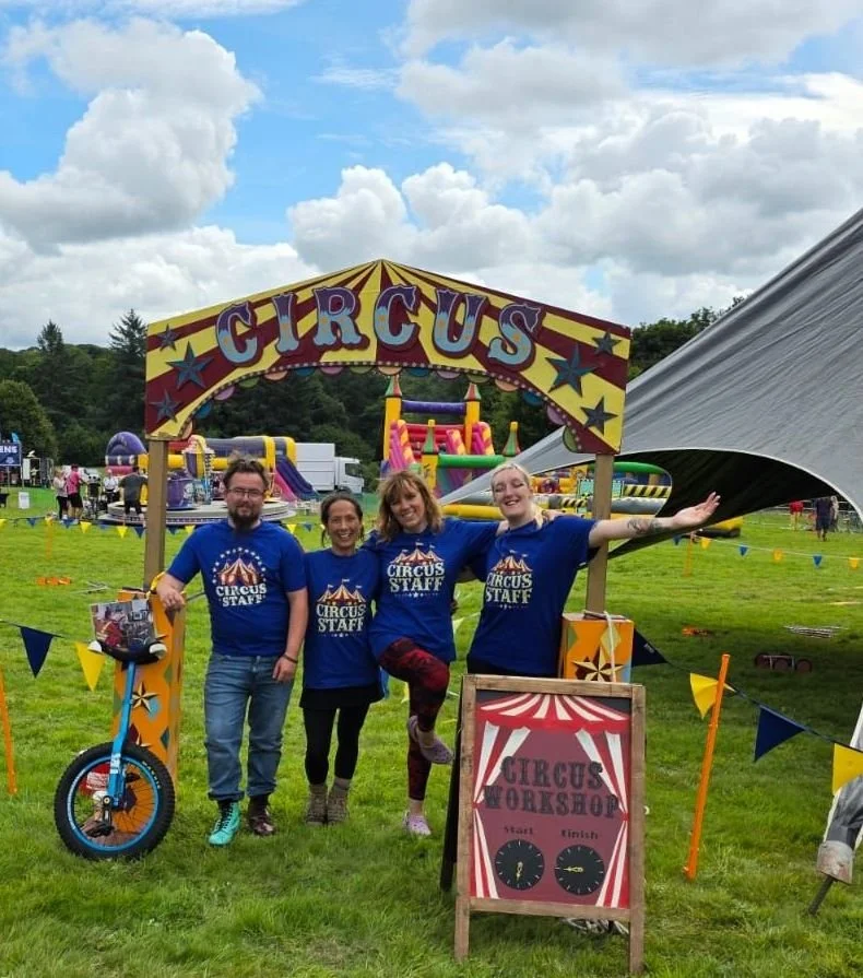 Four people in blue 'Circus Staff' shirts standing under a circus sign at an outdoor event, smiling with a background of colorful inflatable rides and a large tent.
