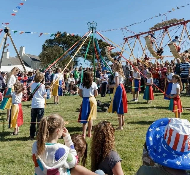 Children and spectators watch a traditional maypole dance at a fairground with a swing ride and flags overhead.