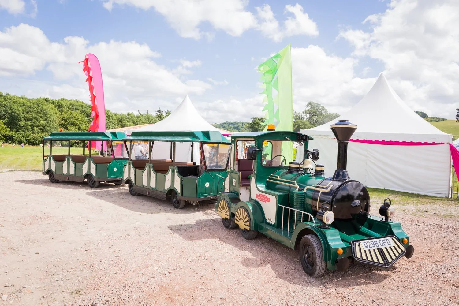 Small green vintage train with open passenger cars and white tents in a park setting under partly cloudy sky.