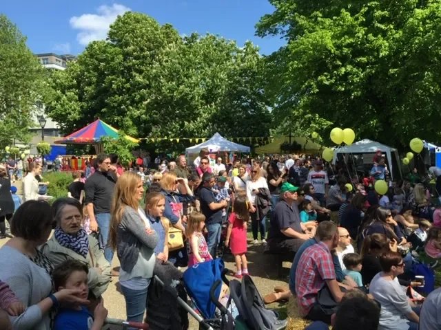 Crowd of people gathered outdoors at a community event with trees, tents, and decorations.