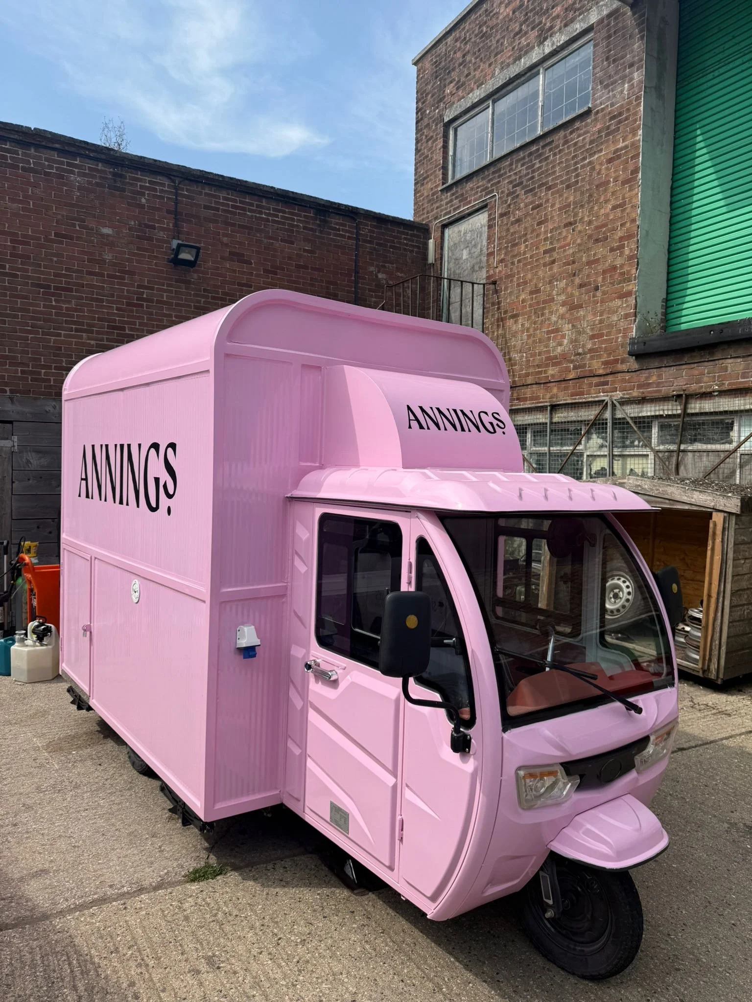 A pink three-wheeled delivery vehicle with the word 'ANNINGS' written on its side and front, parked outdoors on a concrete surface near brick buildings.
