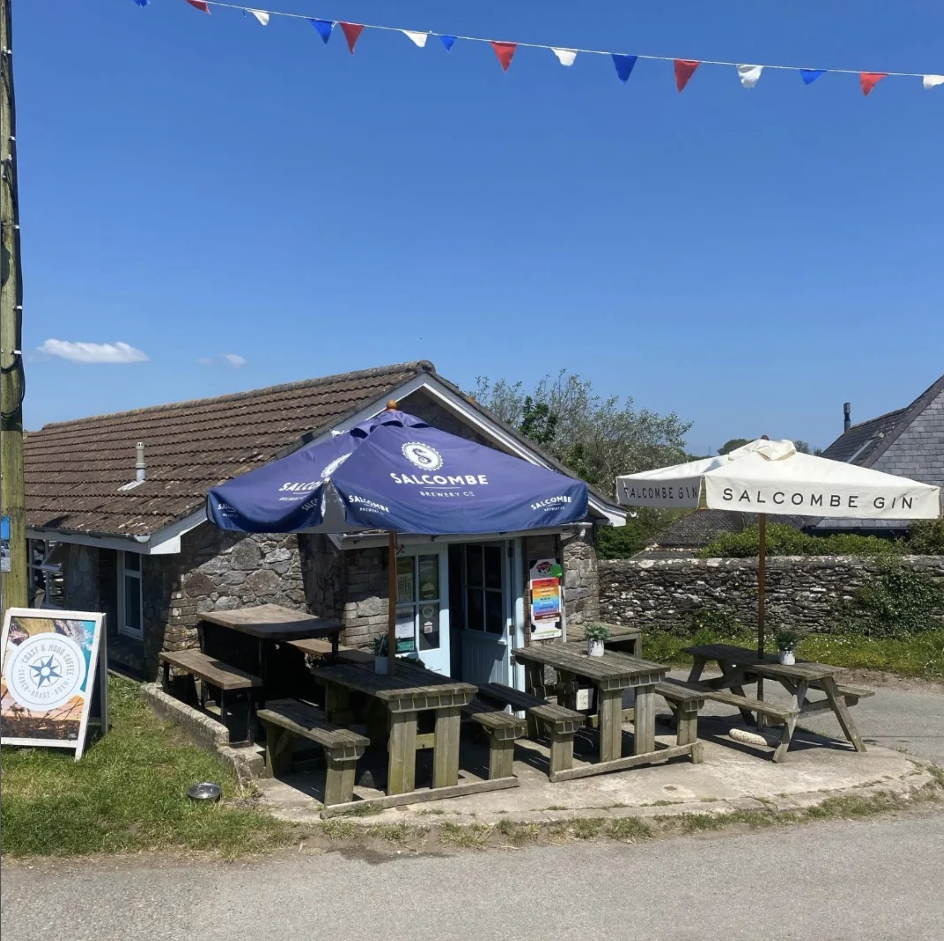 Small stone building with outdoor seating, including wooden picnic tables and benches, shaded by blue and white umbrellas labeled 'Salcombe Gin' and 'Salcombe Brewery'. Red, white, and blue bunting strung across the sky on a sunny day.