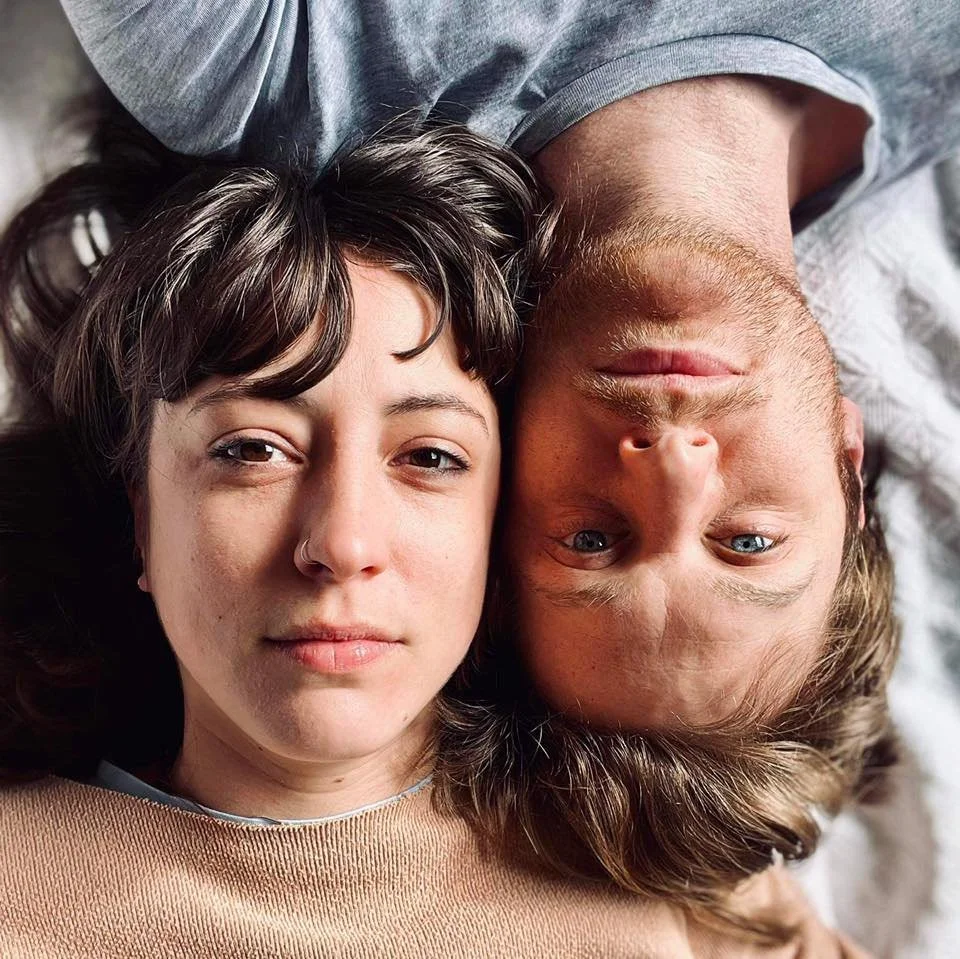 A woman and a man lying on a bed with their heads close together, facing the camera. The woman has dark hair and wears a beige top, while the man has blonde hair and blue eyes.