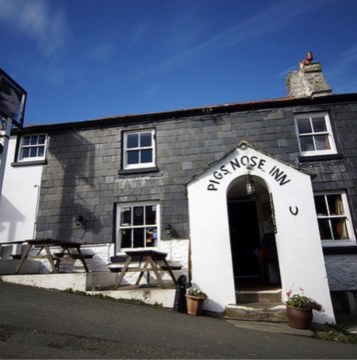 Exterior of the Pig's Nose Inn, a two-story building with gray slate siding, white trimmed windows, and a white entrance archway with the establishment's name. There are picnic tables outside and potted plants near the entrance, under a clear blue sky.