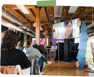 People attending a virtual event or conference in a room with wooden beams, watching a large screen showing a smiling woman on a video call, with holiday decorations including snow-covered trees.