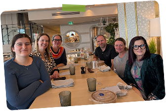 Six people sitting at a long dining table in a restaurant, smiling and posing for the photo. The table has glasses, plates, and a covered dish. The background features decor with plants and a mirror.