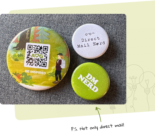 Three round pins with different designs on a gray surface. One pin has a nature scene with a QR code and a man in a hat, with the text "BE INSPIRED." Another pin is white with the text "o-o - Direct Mail Nerd." The third pin is green with the text "DM NERD."