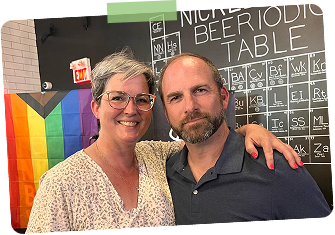 A woman and a man standing together, smiling, at an indoor event with a rainbow pride flag and a periodic table of elements on display behind them.