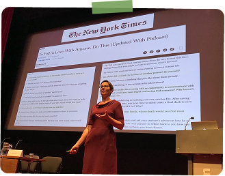 A woman on stage giving a presentation, with a large screen behind her displaying a webpage from The New York Times.