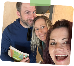 Three smiling people taking a selfie indoors, two women and one man, with furniture and a bookshelf in the background.