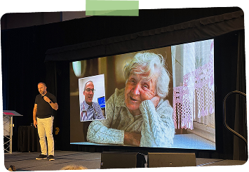 A man stands on stage near a large screen showing a video call with an elderly woman smiling. The scene appears to be in a conference or event setting.