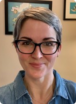 Portrait of a woman with short gray hair, black glasses, and a denim shirt, smiling indoors.