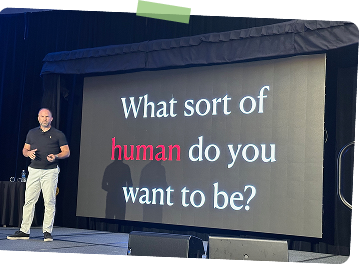 A man standing on stage next to a large screen displaying the question, 'What sort of human do you want to be?'