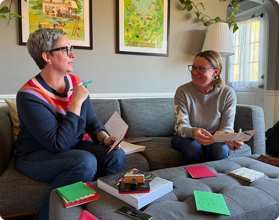 Two women sitting on a gray couch having a conversation with notebooks and papers in their hands, surrounded by colorful papers and books on a coffee table in a living room.