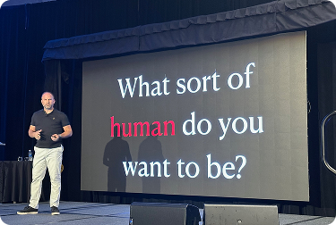 A man standing on a stage next to a large screen displaying the question, 'What sort of human do you want to be?'