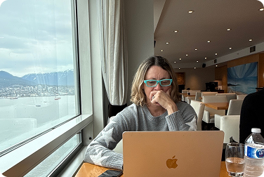A woman with glasses working on a laptop at a restaurant with large windows overlooking a harbor and mountains.