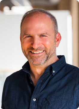 A middle-aged man with a beard and short hair, smiling, wearing a dark blue button-up shirt, in front of a blurred indoor background with sunlight.