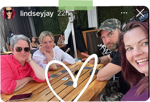 Four people sitting around a table outdoors, smiling for a selfie, with a heart shape drawn in the foreground.