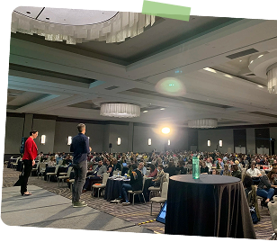 Large conference room filled with people seated at tables, with a few attendees standing and speaking on stage. Overhead lighting and chandeliers illuminate the room.