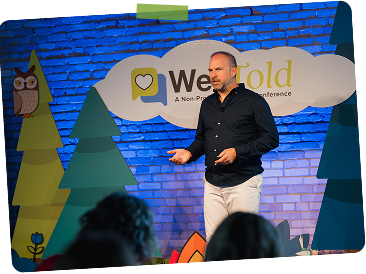 A man standing on a stage giving a presentation at a conference. The backdrop includes decorative trees, an owl, and a banner that reads 'We Gold' and 'A Non-P...
