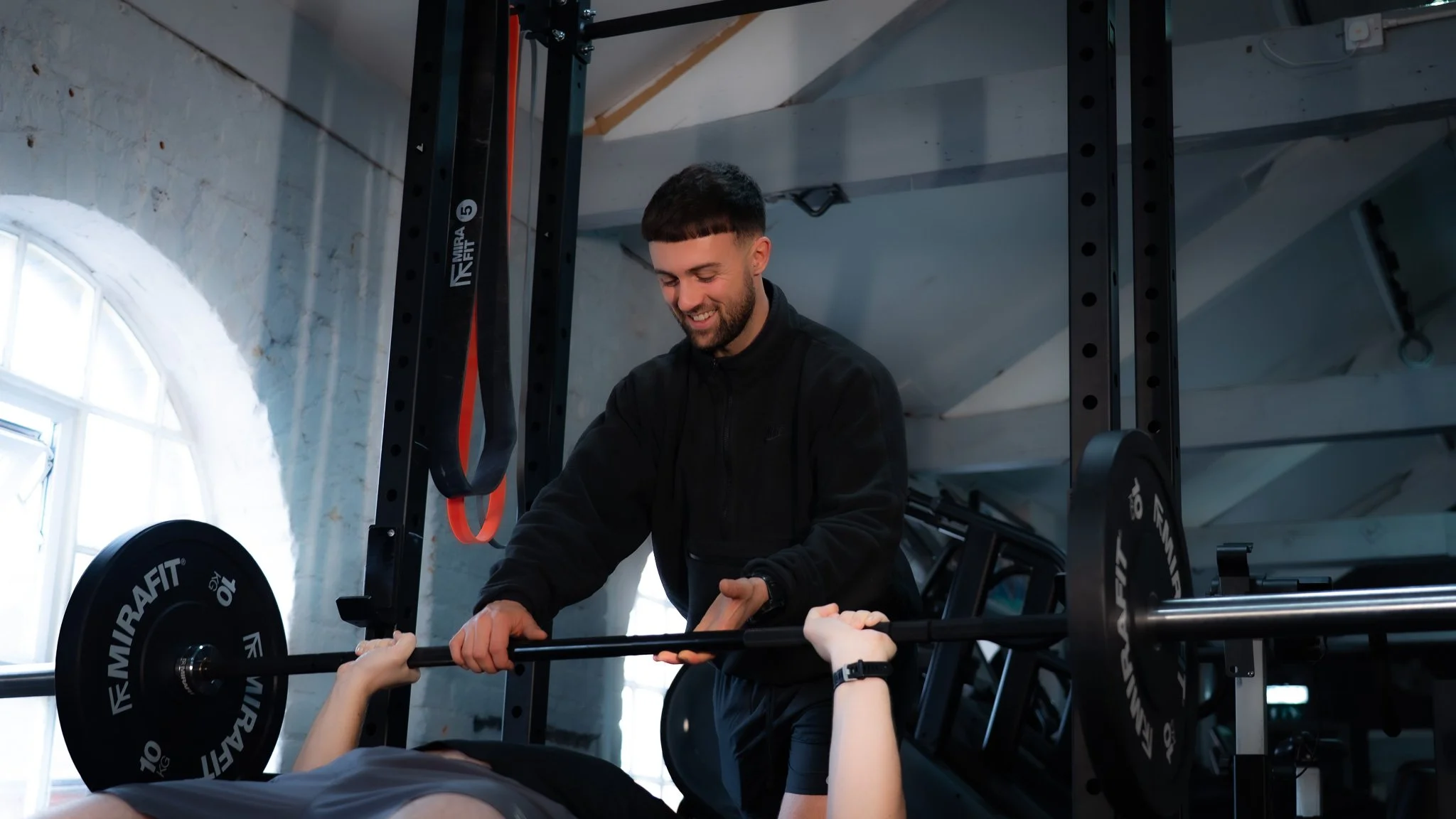 A man smiling at a woman who is lying on a bench press, lifting a barbell in a gym with bright natural light coming through windows.