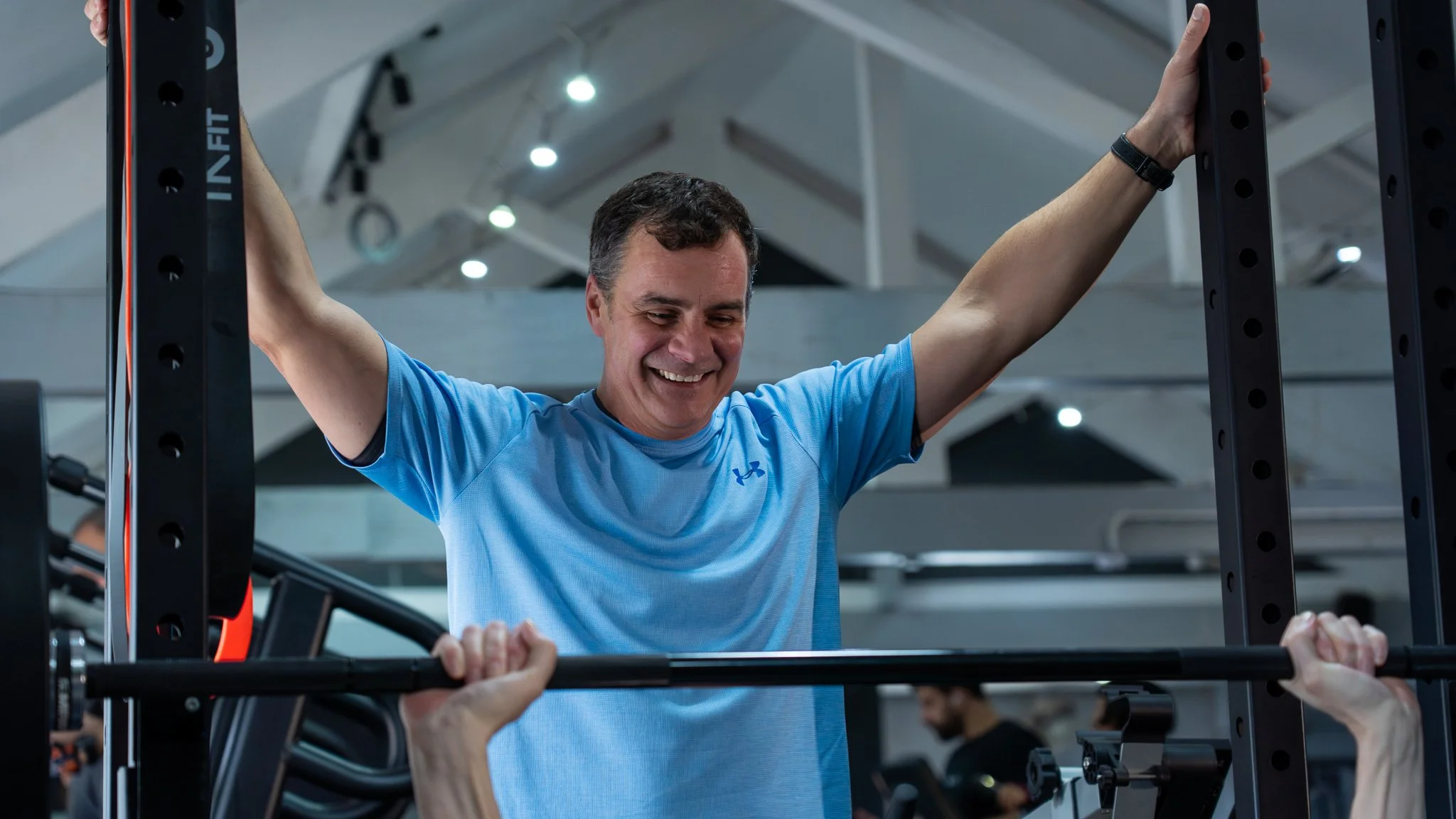 A man in a blue athletic shirt smiling while lifting a barbell during a workout in a gym.